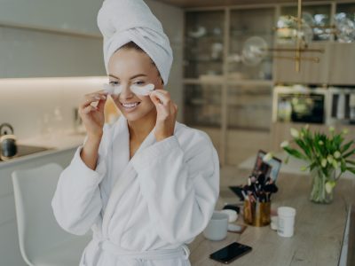 Young happy healthy woman in bathrobe and towel turban on head taking off collagen hydrogel beauty patches and smiling while doing beauty or cosmetic procedures at home. Personal care concept